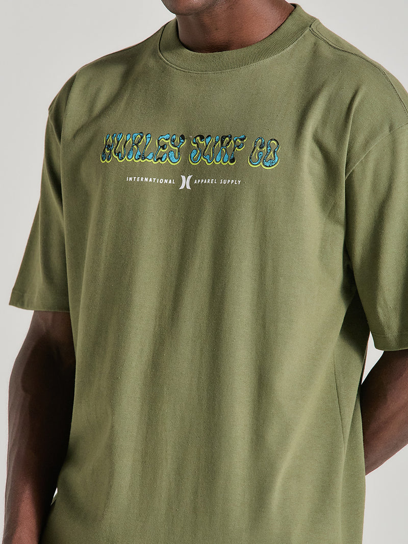Close da camiseta de algodão oversized verde militar manga curta com estampa "Hurley Surf Co." em lettering estilizado e fontes adicionais abaixo.