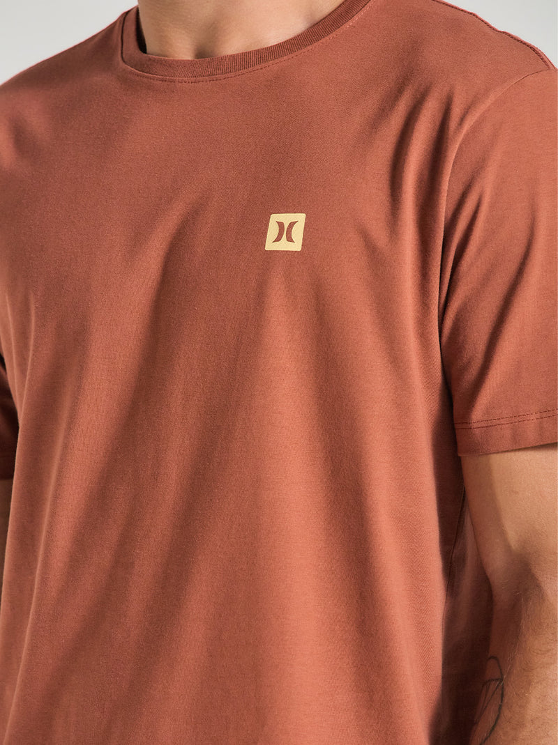 Homem vestindo camiseta em algodão marrom com gola redonda, mangas curtas e logotipo pequeno em amarelo no lado esquerdo do peito.