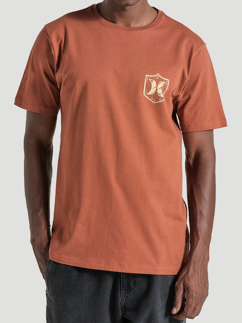 Homem vestindo camiseta marrom manga curta estampa no lado esquerdo do peito, em formato de escudo com detalhes em bege e o logotipo "Hurley".