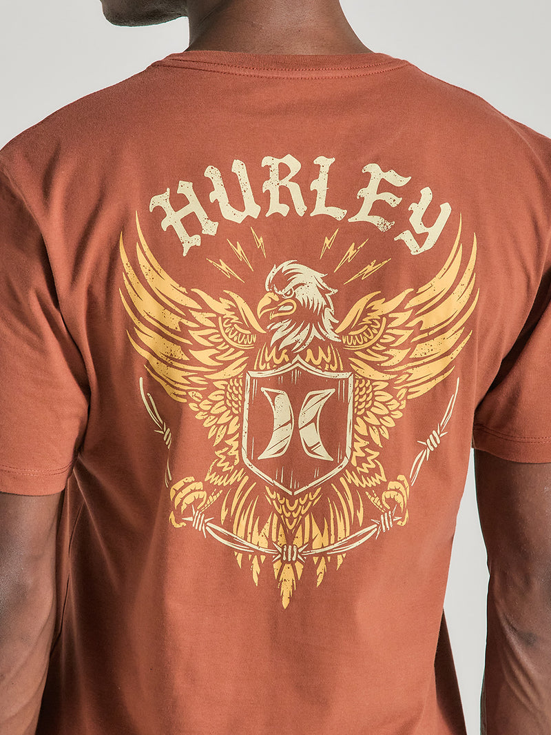 Camiseta marrom de manga curta estampa nas costas, apresentando uma águia dourada estilizada, escudo central com o logotipo "Hurley" acima.