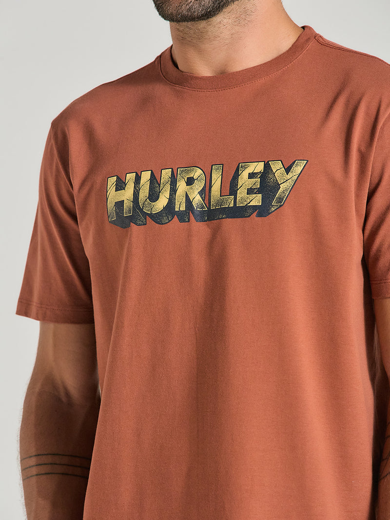 Homem usando camiseta marrom manga curta estampada com estampa frontal do logotipo "Hurley", o estilo é do tipo desgastado nas cores amarela e preta.
