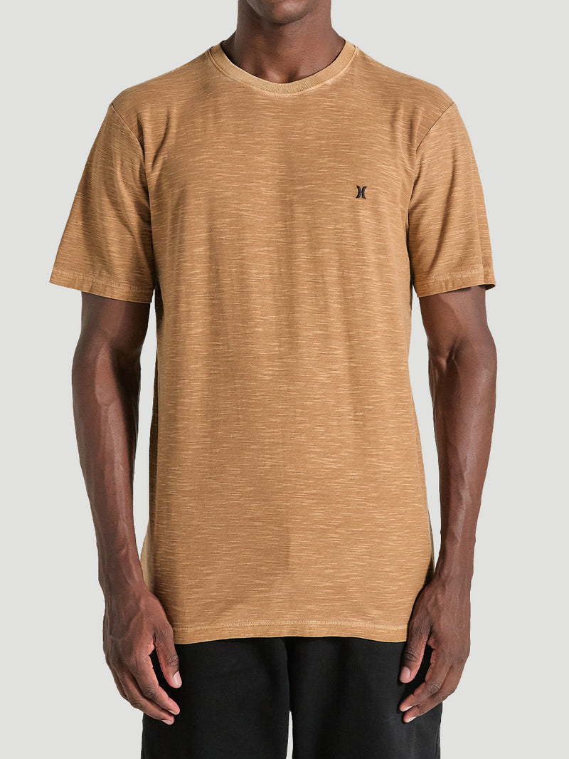 Homem vestindo camiseta marrom tinturada manga curta com pequeno logo no peito, combinada com bermuda preta. Fundo cinza claro.