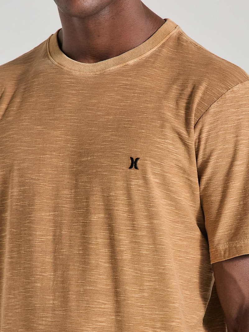 Close da camiseta marrom tinturada manga curta com tecido texturizado e pequeno logo preto bordado no peito. Fundo neutro claro.