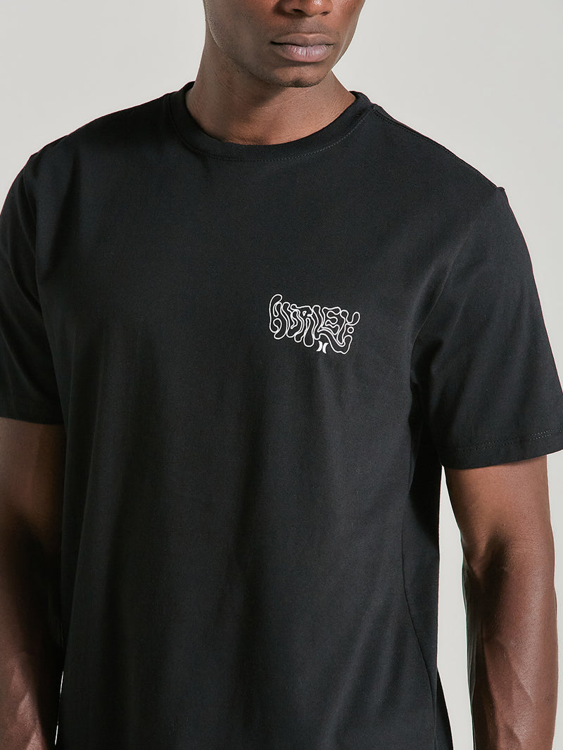 Homem veste uma camiseta preta manga curta estampa relevo com arte branca no lado esquerdo do peito, exibindo um design abstrato e estilizado.