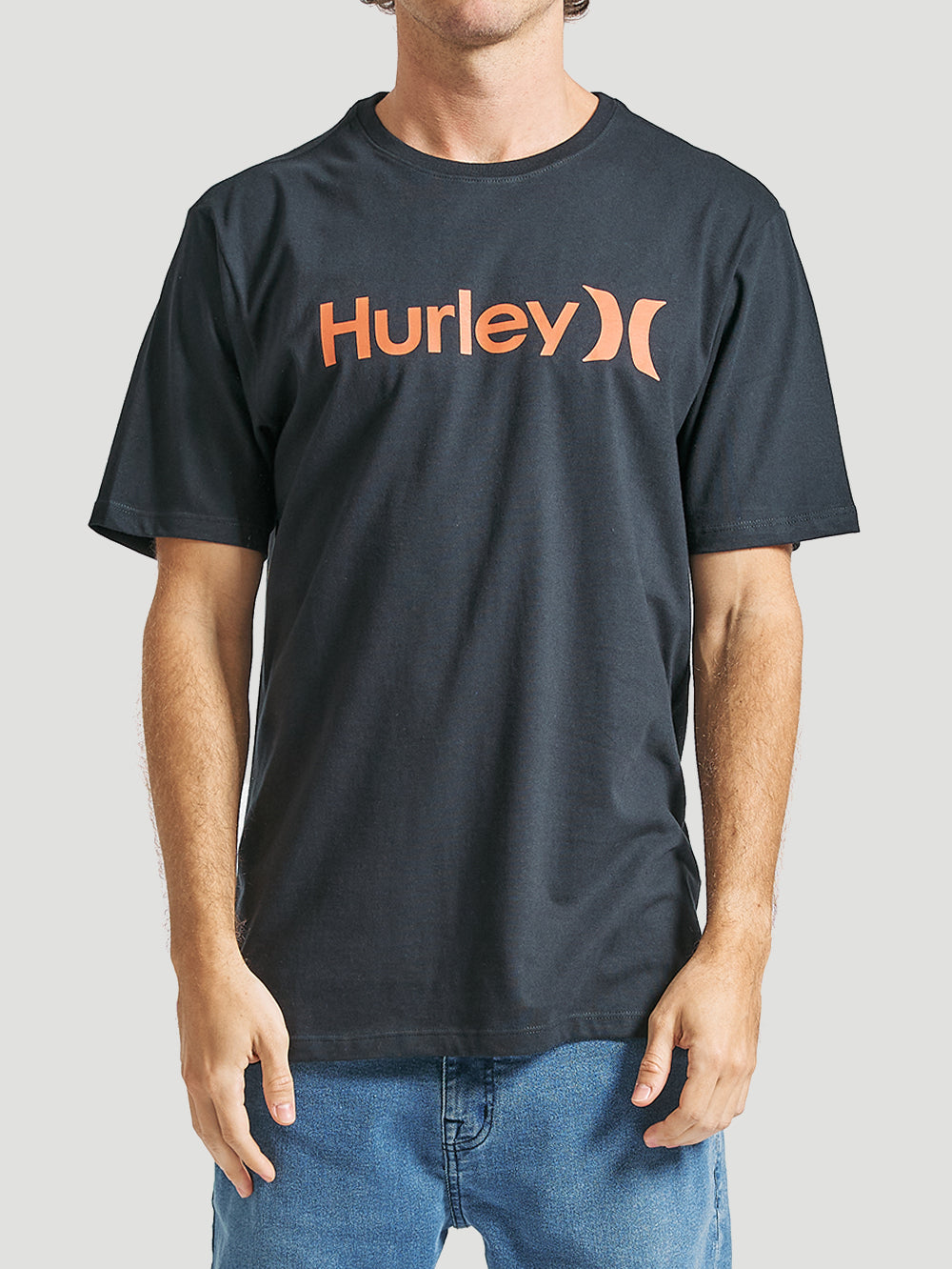 Homem veste uma camiseta preta manga curta com estampa Hurley frontal em laranja, combinada com jeans azul claro, em fundo neutro.