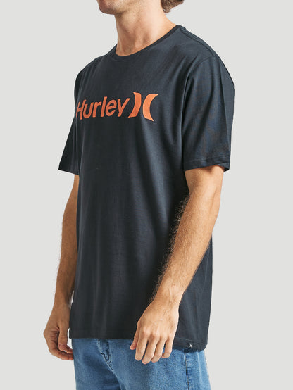 Homem veste uma camiseta preta manga curta com estampa frontal laranja Hurley, tecido liso e caimento reto, combinada com jeans azul, em fundo claro.