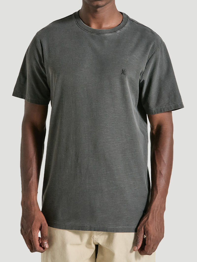 Camiseta preta tinturada manga curta com pequeno logo no peito, combinada com bermuda bege. Fundo cinza claro.