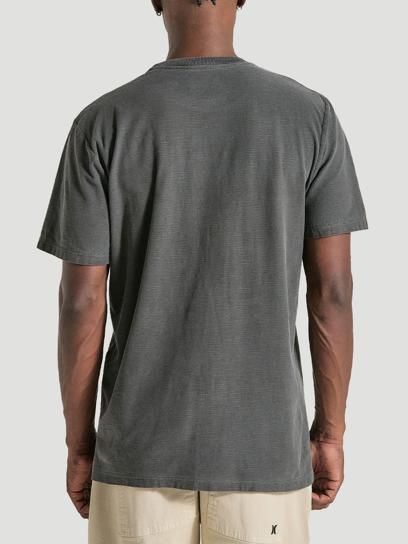Camiseta preta tinturada manga curta vista de costas, combinada com bermuda bege clara. Fundo cinza claro.