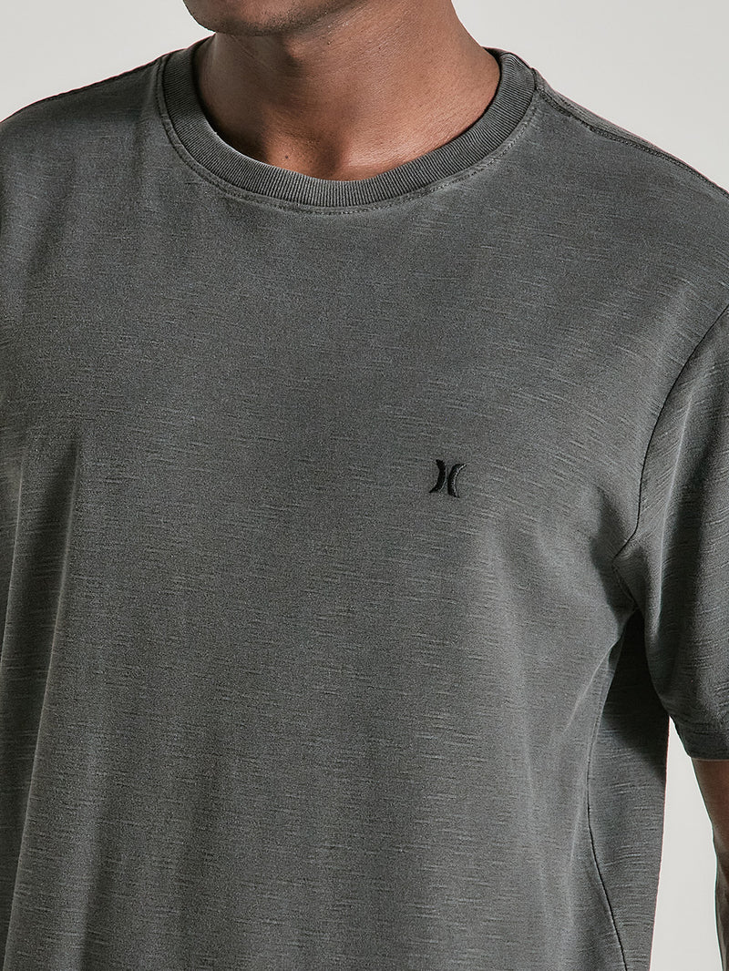 Homem vestindo camiseta preta tinturada manga curta com textura sutil e pequeno logo bordado no peito em preto. Fundo claro e neutro.