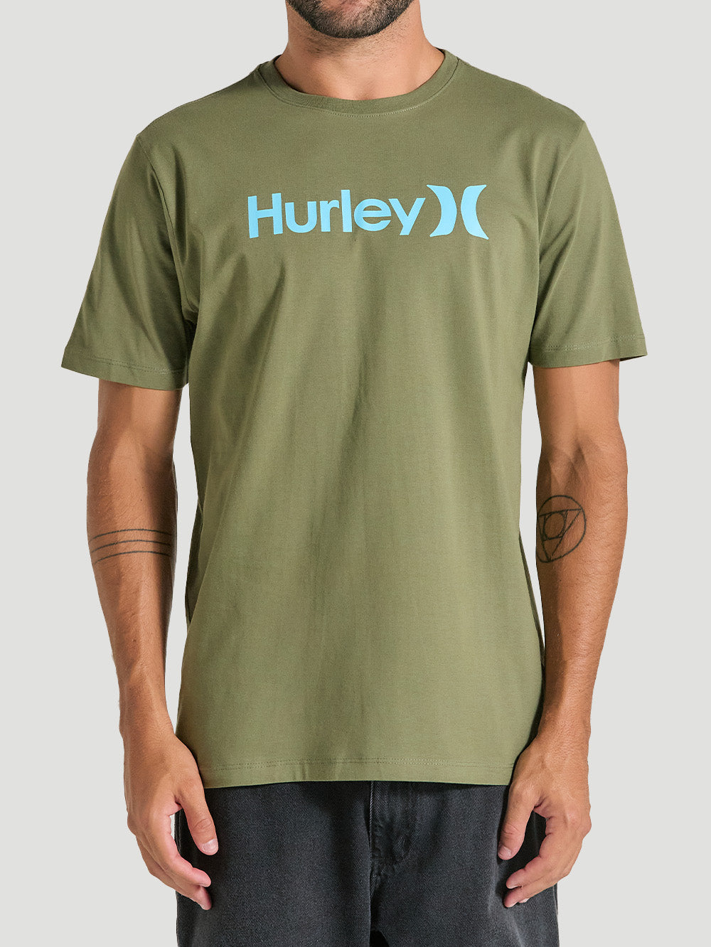 Homem veste uma camiseta verde manga curta com estampa frontal azul Hurley, de tecido liso e caimento reto, combinada com jeans preto, em fundo neutro.