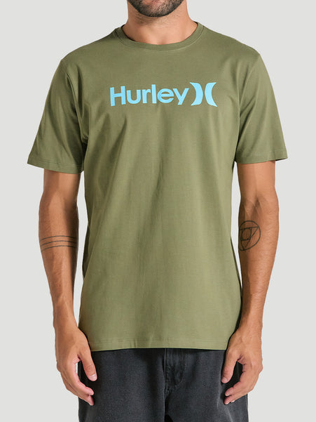 Homem veste uma camiseta verde manga curta com estampa frontal azul Hurley, de tecido liso e caimento reto, combinada com jeans preto, em fundo neutro.