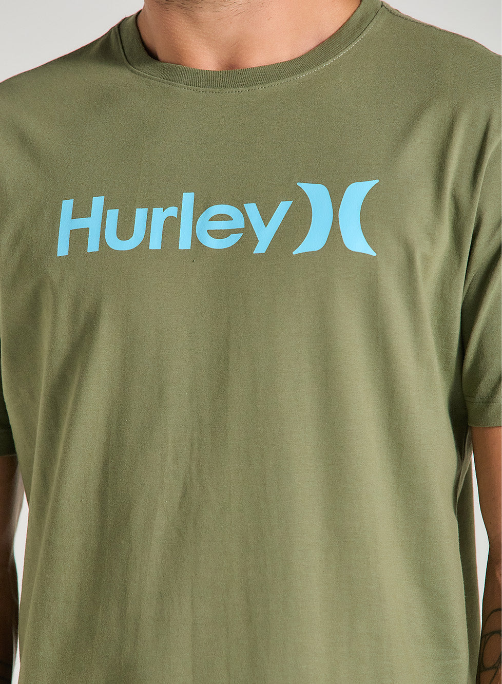 Homem veste uma camiseta verde manga curta com estampa frontal azul claro Hurley, de tecido liso e caimento reto, em fundo claro.