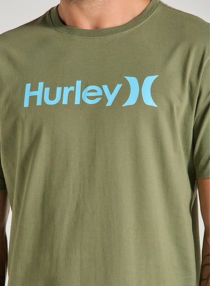 Homem veste uma camiseta verde manga curta com estampa frontal azul claro Hurley, de tecido liso e caimento reto, em fundo claro.