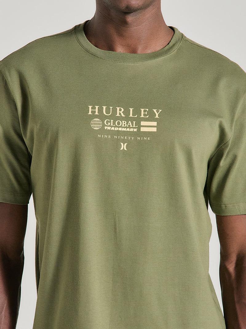 Homem vestindo camiseta verde militar estampa frontal manga curta com escrita em bege, logotipo e texto, fundo neutro e foco na parte superior do torso.