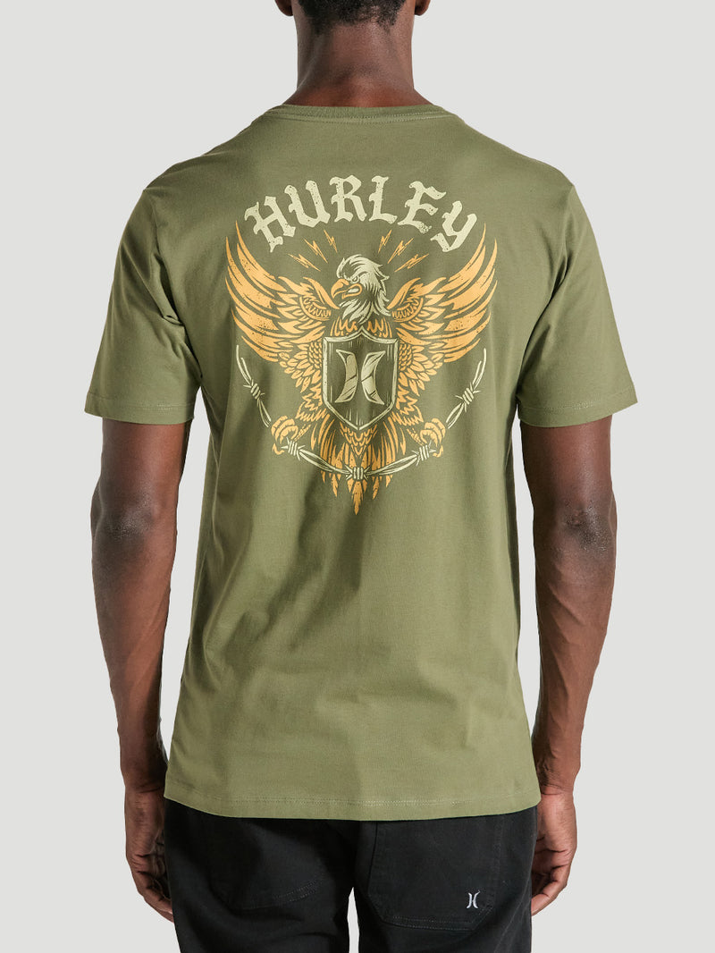 Homem vestindo camiseta verde militar manga curta estampa nas costas, apresentando uma águia dourada com asas abertas, escudo central e o texto "Hurley".