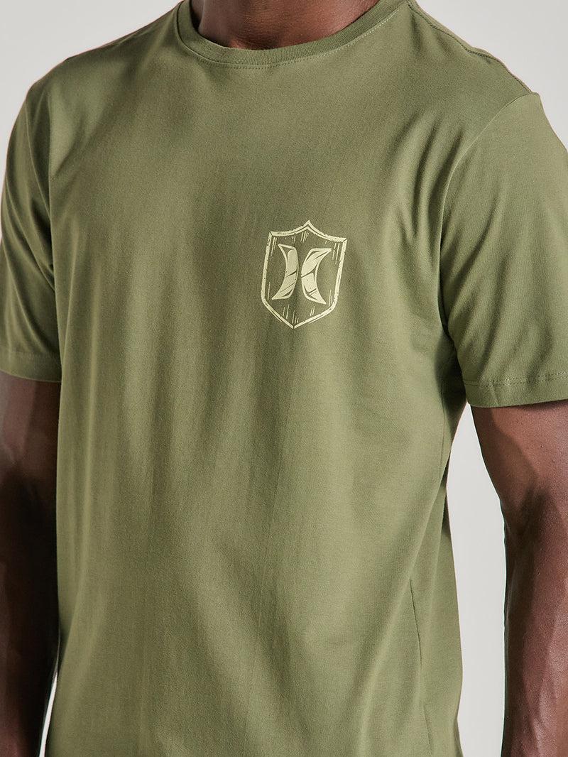 Camiseta verde militar manga curta estampa discreta no lado esquerdo do peito, em formato de escudo bege com elemento estilizado ao centro.
