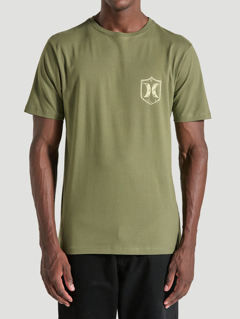 Homem vestindo camiseta verde militar manga curta estampa pequena no lado esquerdo do peito, apresentando um escudo estilizado em bege com símbolo central.