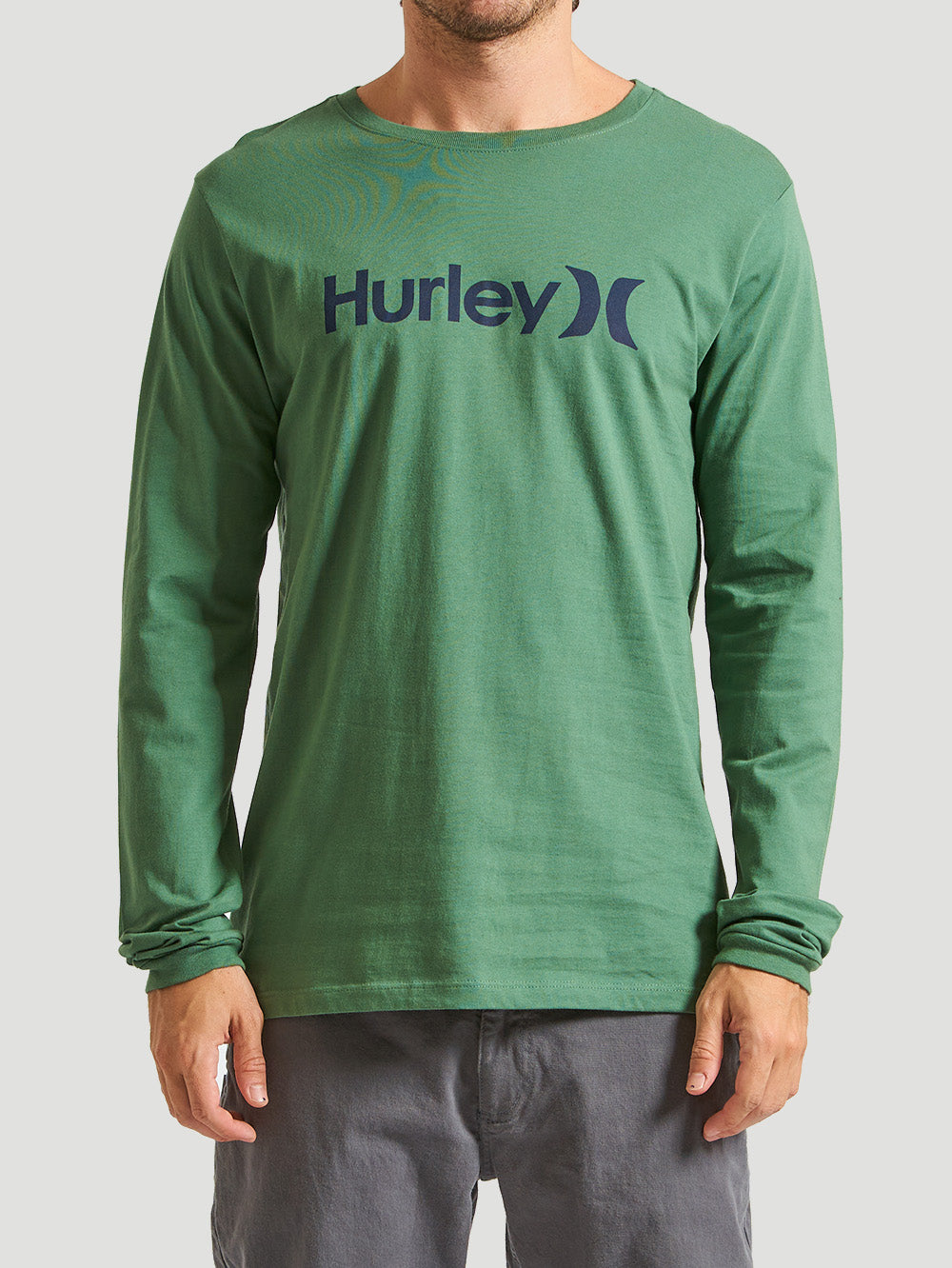 Camiseta verde militar manga longa com estampa do logo "Hurley" em azul escuro, combinada com calça cinza. Fundo cinza claro.