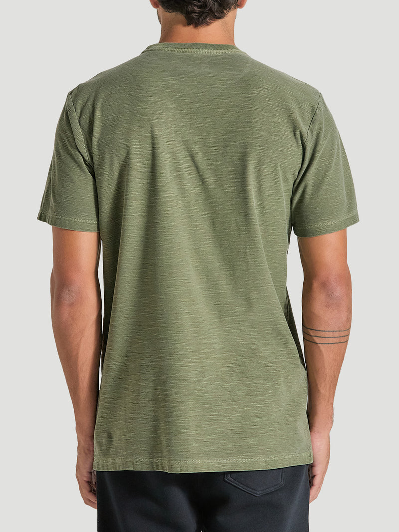 Camiseta verde militar tinturada manga curta vista de costas, com tecido de textura leve. Combinada com bermuda escura. Fundo cinza claro.