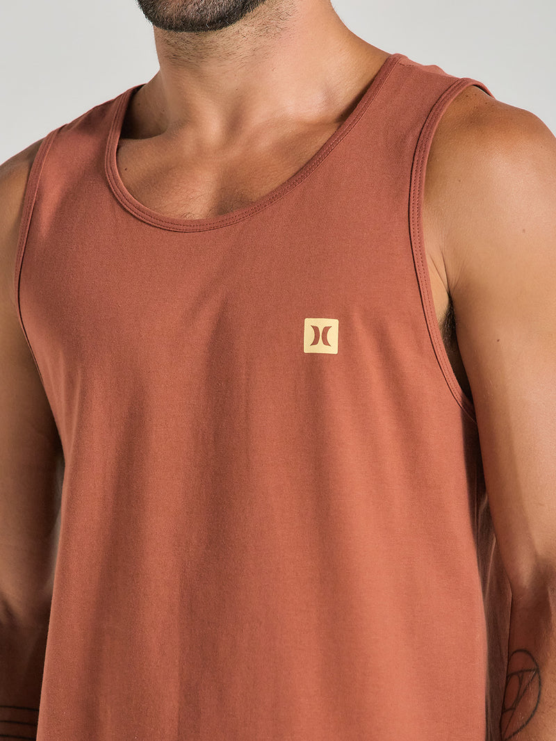 Homem vestindo regata marrom esportiva com logo discreto em amarelo no lado esquerdo do peito. A peça possui alças largas e tecido leve.
