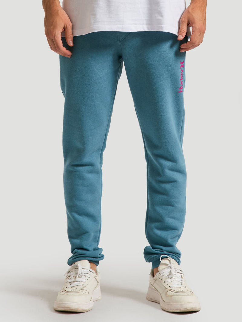Homem veste uma calça moletom azul básica ajustada com leve elasticidade, combinada com tênis brancos e camiseta branca, em fundo neutro.