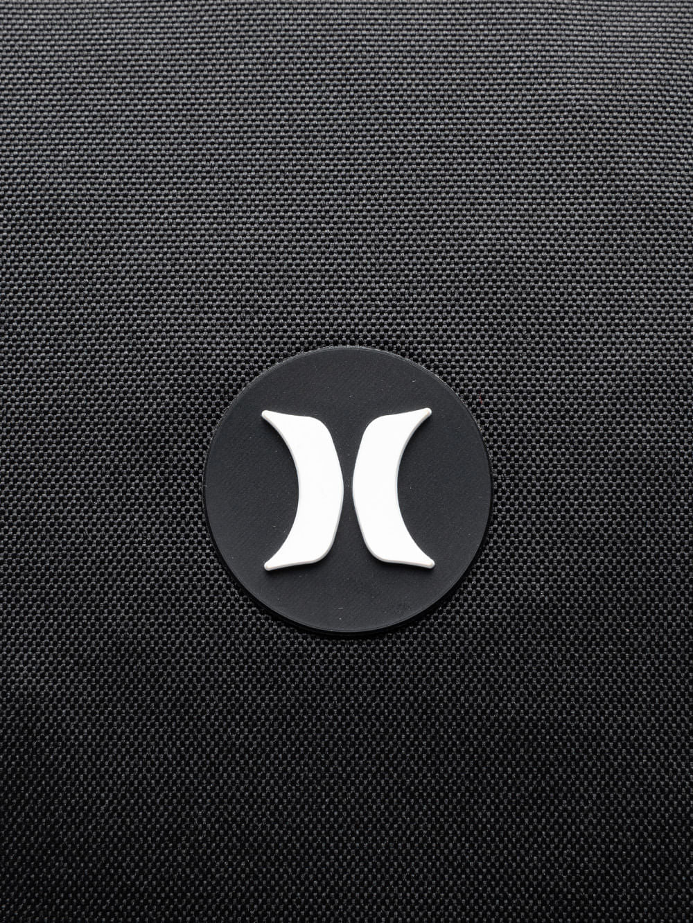 Close de logo branco na mochila Hurley em destaque no tecido preto texturizado. Design minimalista e moderno.