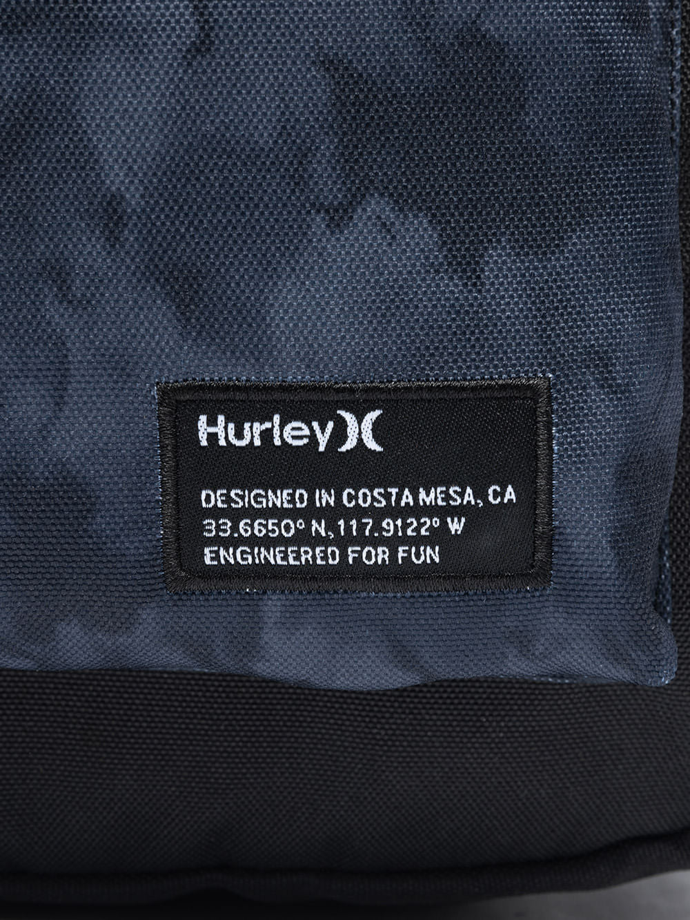 Detalhe da mochila Hurley com etiqueta preta contendo o logotipo branco, coordenadas geográficas e a frase "Engineered for fun" impressa.