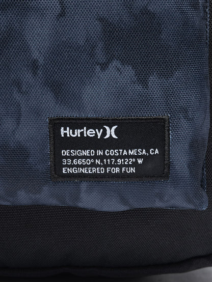 Detalhe da mochila Hurley com etiqueta preta contendo o logotipo branco, coordenadas geográficas e a frase "Engineered for fun" impressa.